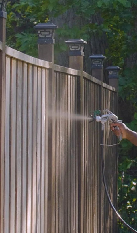 Sprayer applying stain to vertical wood fence pickets with decorative post caps in Edina, MN showing mid-application on the yard-facing side