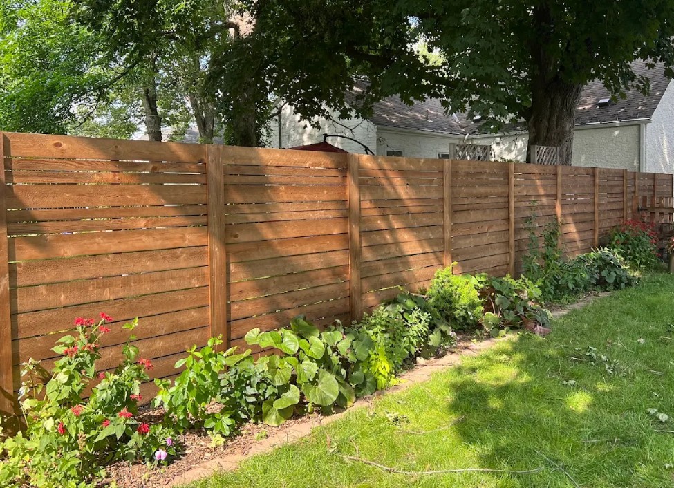 Freshly stained horizontal slat wood privacy fence in an Edina, MN backyard with rich honey-brown color, lush green landscaping, and red flowers along the base
